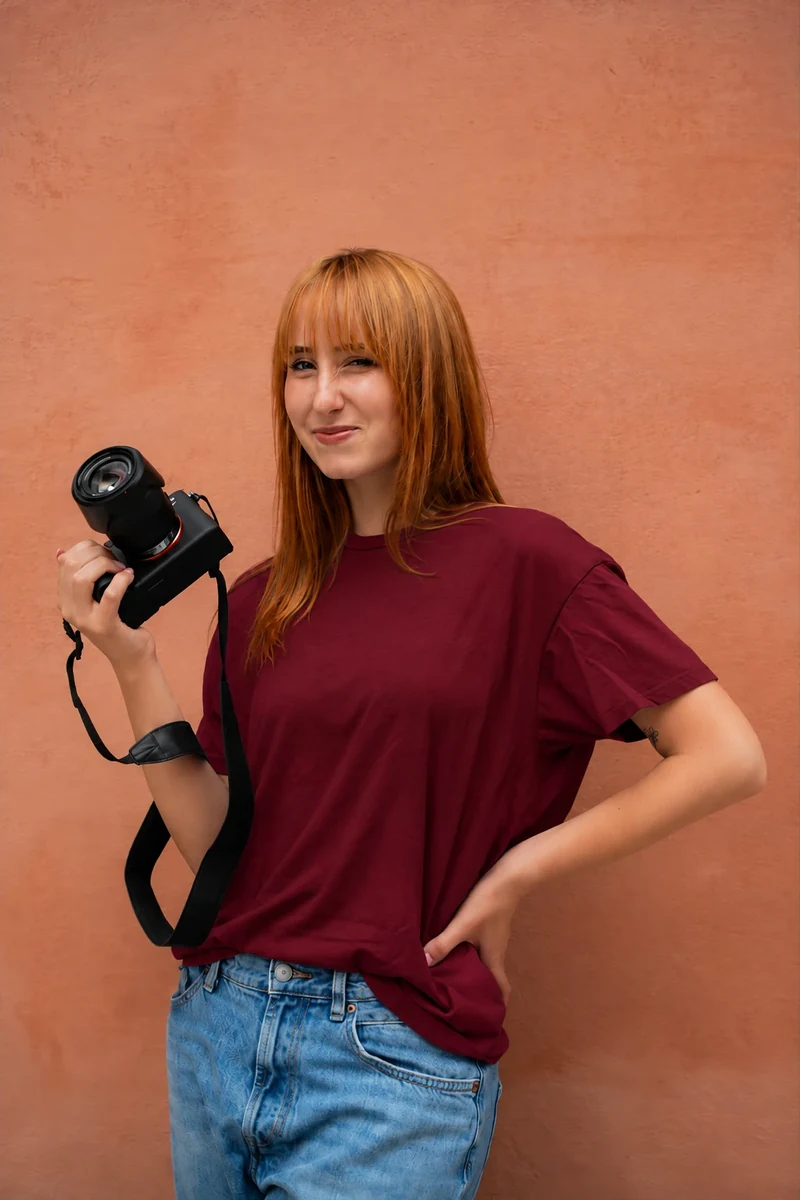 Margherita Giorgi con la maglia bordeaux e la macchina fotografica in mano, davanti a un muro color terracotta