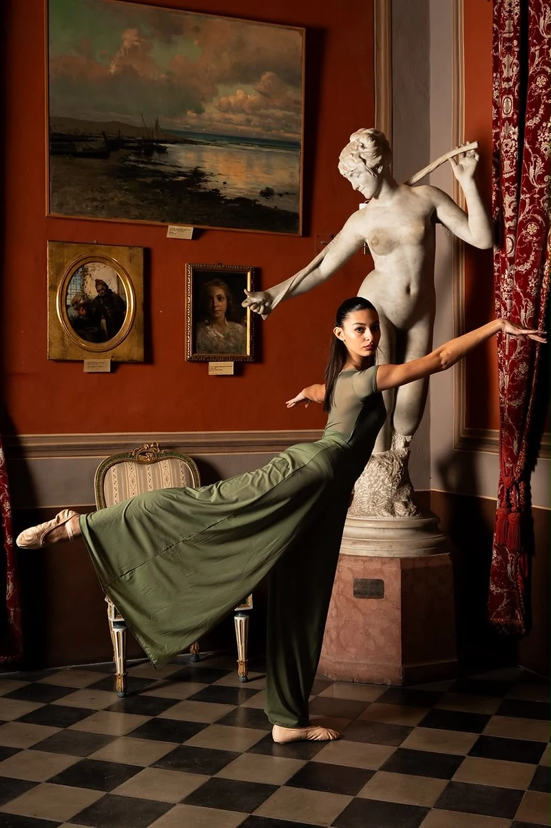 Ballerina in abito verde in arabesque accanto a una statua, su pavimento a scacchi