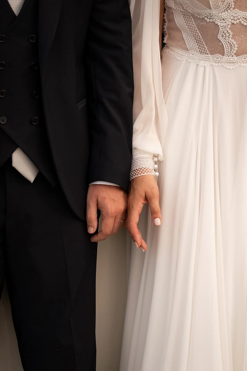 Sposi uno accanto all'altra che si sfiorano la mano, abito in tulle e tight scuro
