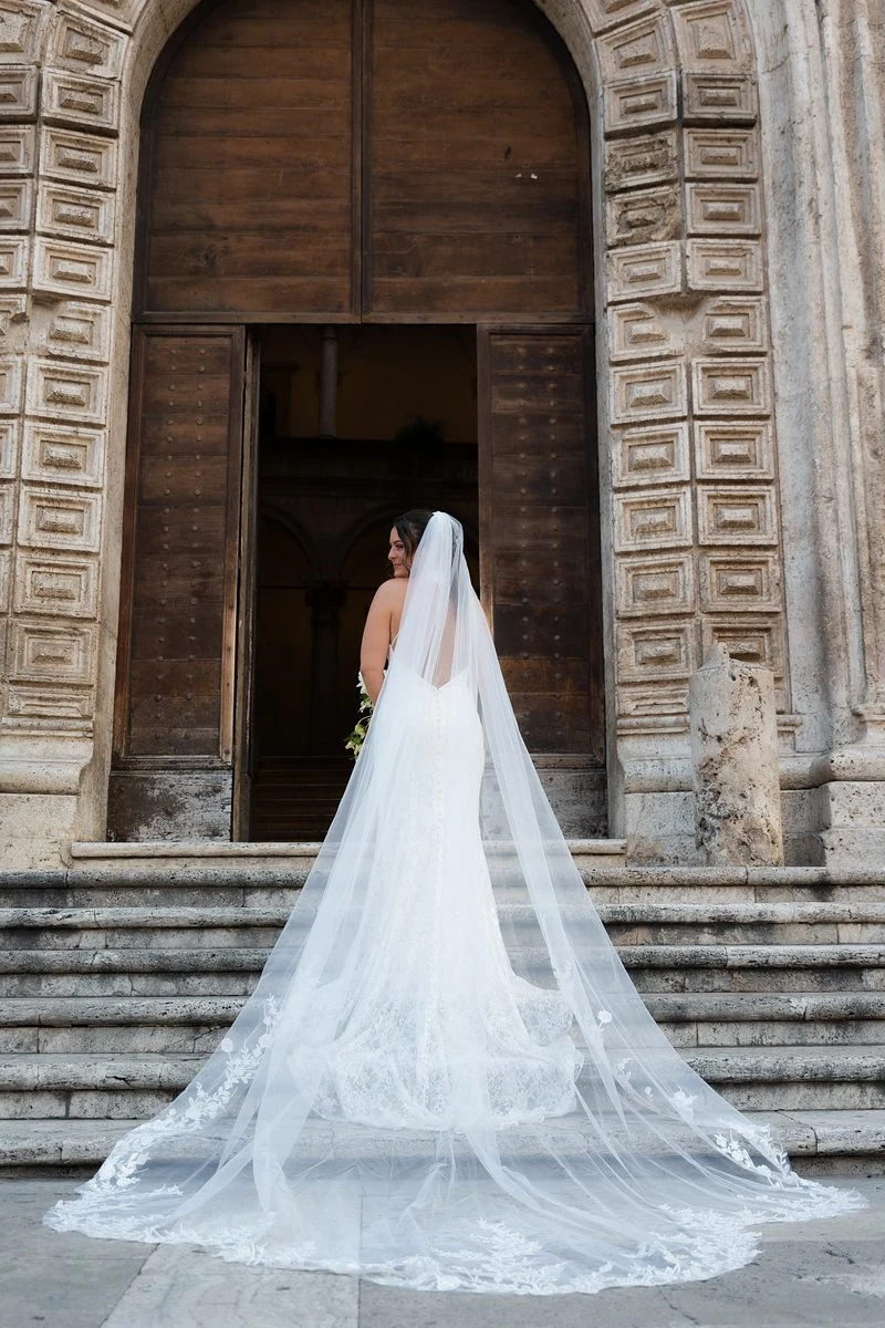 Sposa di spalle con il velo lungo sulla scalinata davanti al portone di una chiesa