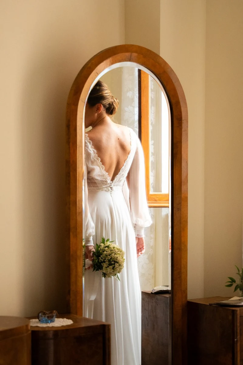 Sposa con il bouquet che si guarda nello specchio ad arco, di spalle