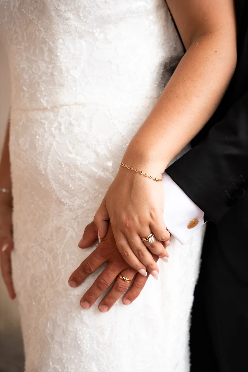 Mano dello sposo che stringe quella della sposa sull'abito di pizzo, con le fedi
