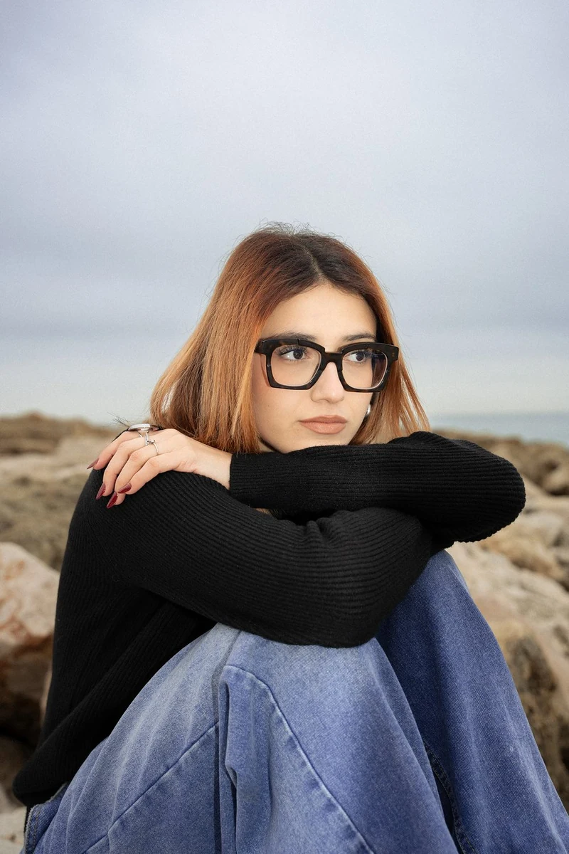 Ragazza con gli occhiali seduta sugli scogli, abbracciata alle ginocchia
