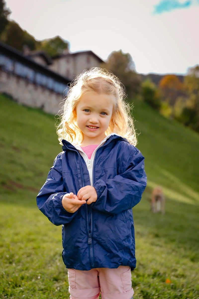 Bambina con la giacca blu in piedi su un prato, capelli biondi in controluce